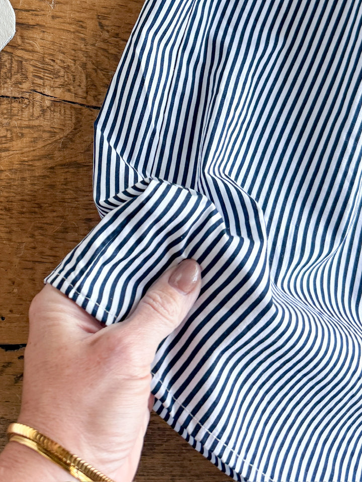 A hand holds the edge of The Tererro, a blue and white pinstripe midi dress by Savannah Sevens western life{&}style, against a rustic wooden surface.