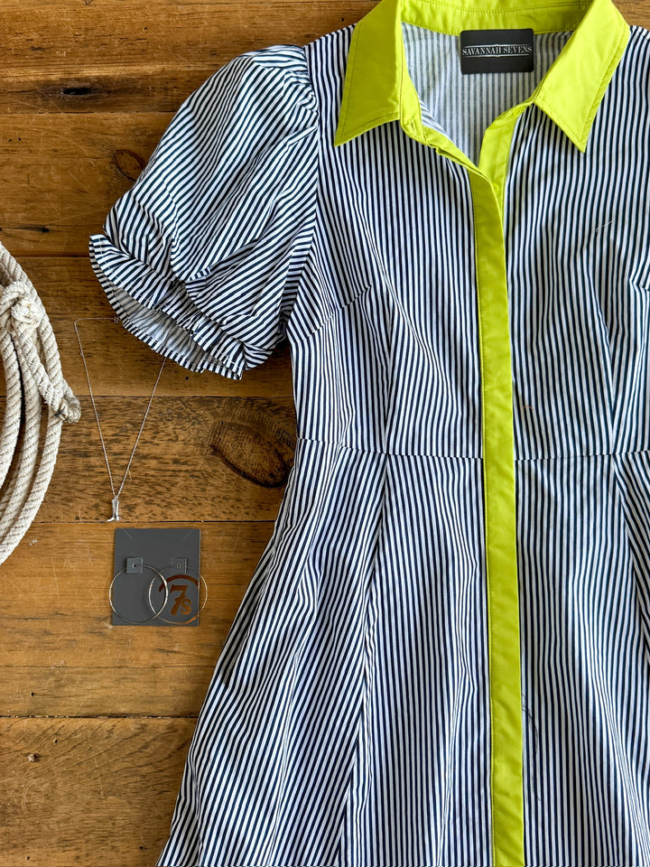 The Tererro by Savannah Sevens western life{&}style, a black-and-white pinstripe midi dress with short sleeves and a bright yellow collar and trim, is displayed on a wooden surface beside a silver pendant necklace.