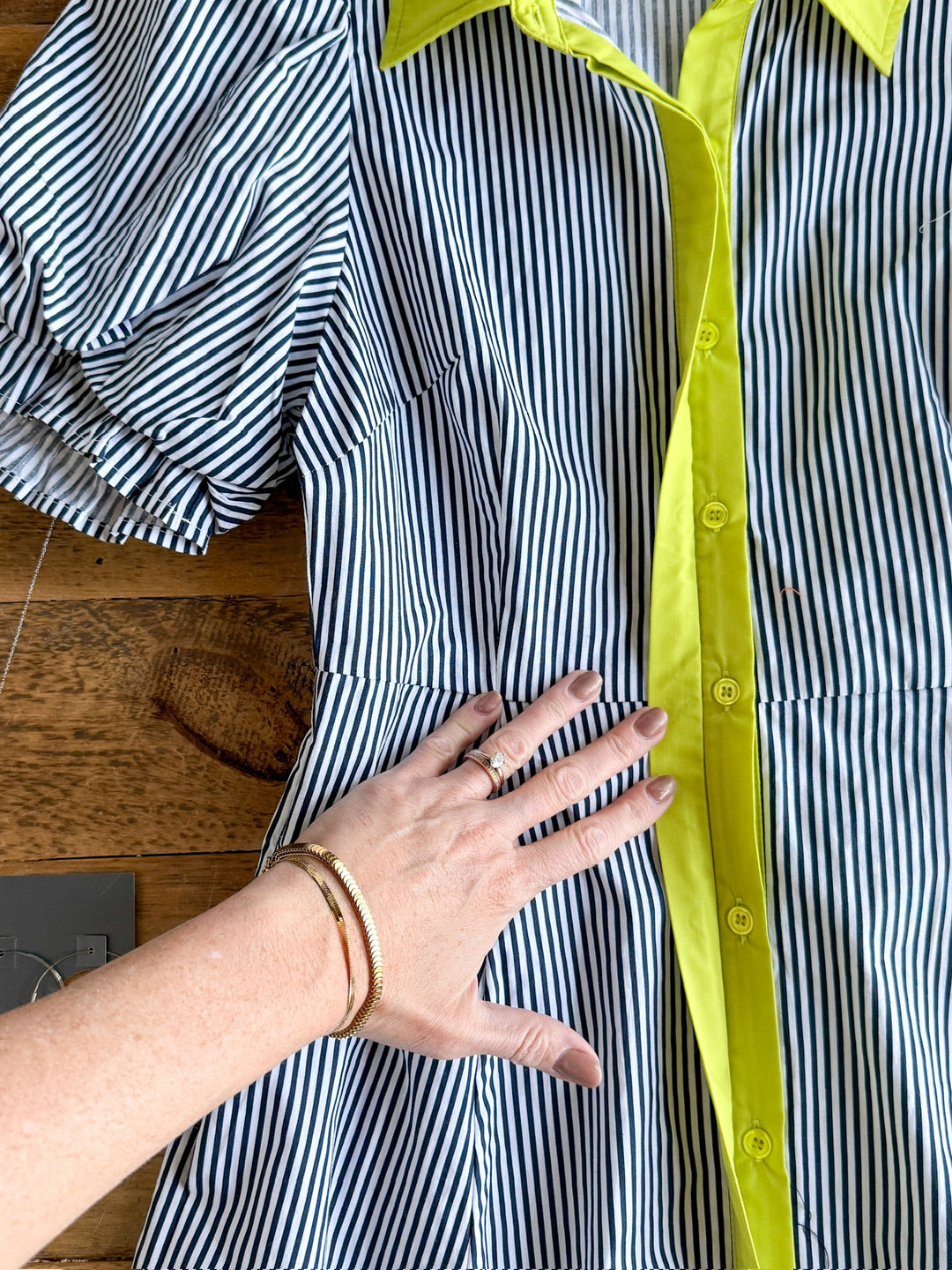 A hand with rings and a bracelet rests on The Tererro midi dress by Savannah Sevens western life{&}style, featuring blue-and-white pinstripes, bright yellow buttons, collar, and trim, laid out on a wooden surface.