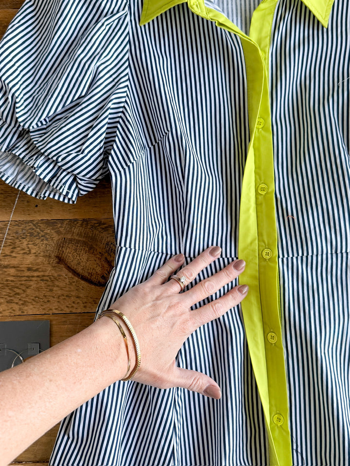A hand with rings and a bracelet rests on The Tererro midi dress by Savannah Sevens western life{&}style, featuring blue-and-white pinstripes, bright yellow buttons, collar, and trim, laid out on a wooden surface.