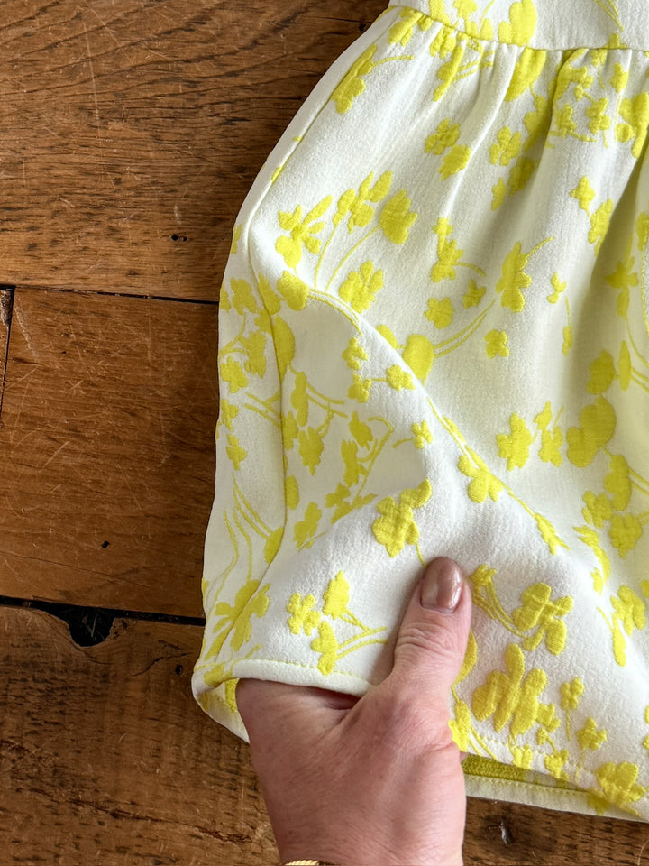 A hand holds the hem of The Spring Valley white fabric with an electric lemon floral pattern by Savannah Sevens western life{&}style, set against a wooden surface.