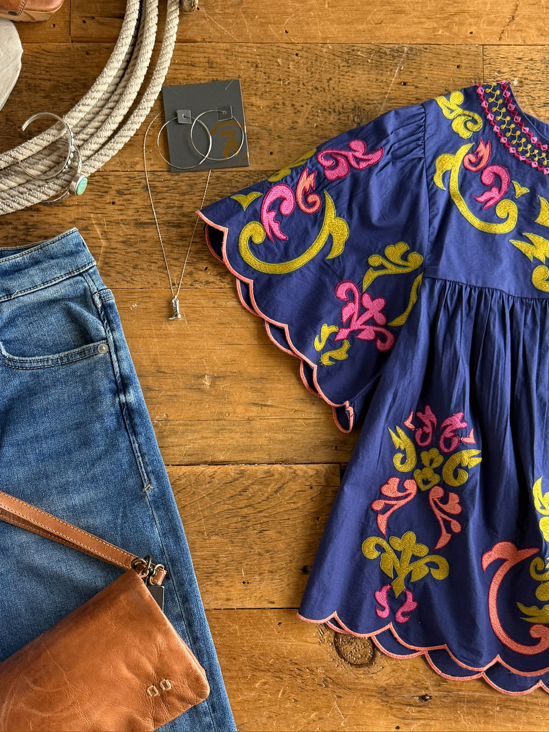 Flat lay of The Montevello navy flutter sleeve blouse by Savannah Sevens western life{&}style, featuring colorful floral embroidery, paired with blue jeans, a brown leather purse, silver necklace, hoop earrings, and coiled rope on wood.