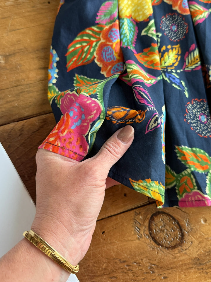 A hand shows a gold bracelet holds the edge of The Pacifica cotton floral top by Savannah Sevens western life{&}style, featuring vibrant patterns, against a wooden background.