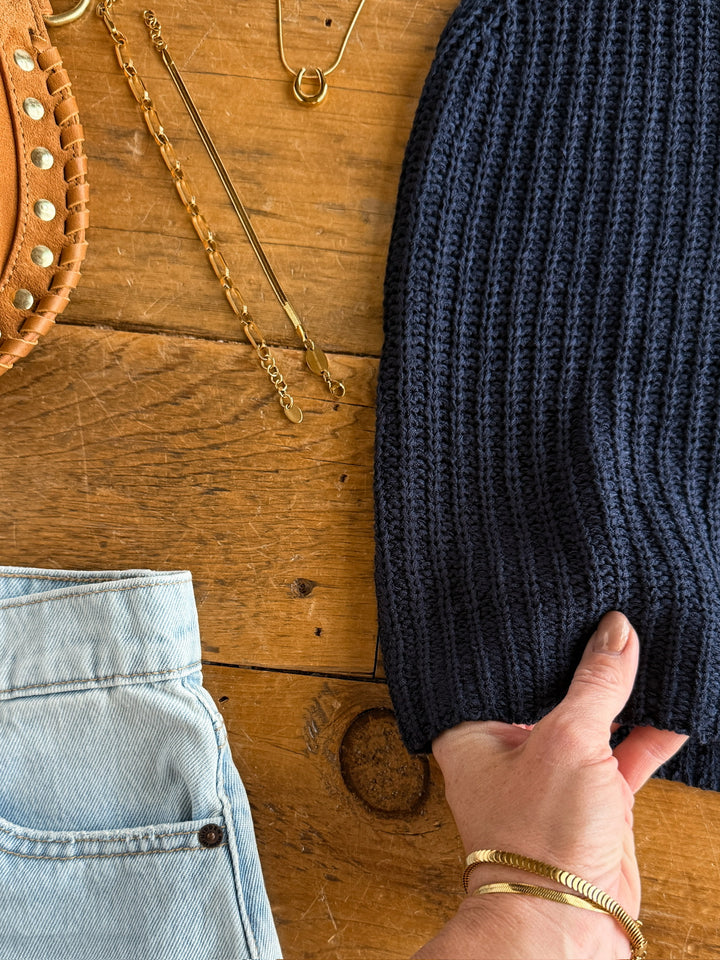 A hand holds the ribbed hem of The Talco navy knit top by Savannah Sevens western life{&}style, displayed on wood beside light blue jeans, a tan studded bag, and gold jewelry.
