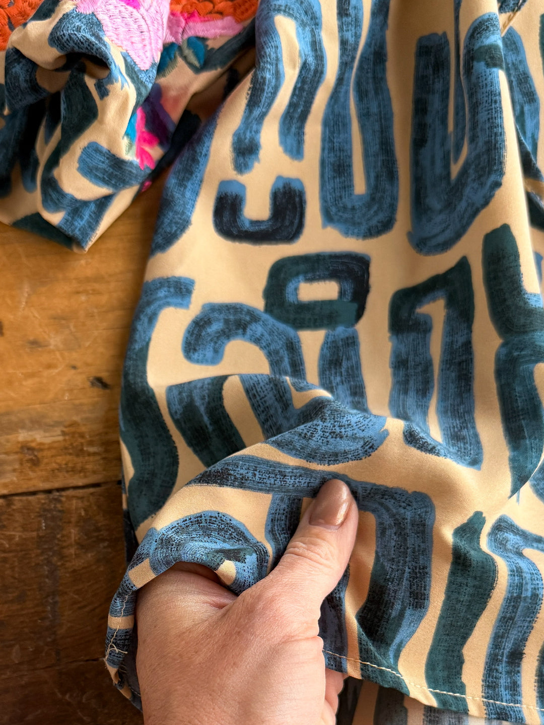A hand  holds a beige fabric featuring bold blue patterns on wood. At the top, The Flora floral embroidered top by Savannah Sevens western life{&}style showcases bright pink and orange details.