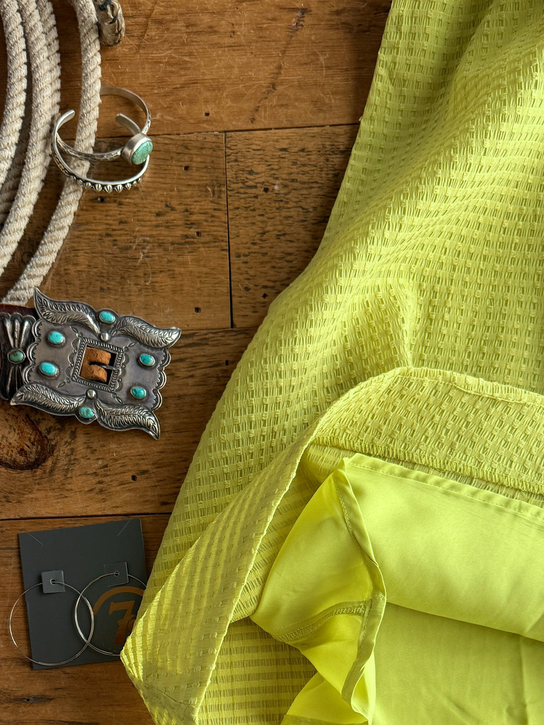 The Kalewa by Savannah Sevens western life{&}style—a yellow textured tri-blend with a ruched halter neckline—lies on wood next to a silver turquoise-studded belt buckle, turquoise bracelet, coiled rope, and hoop earrings.