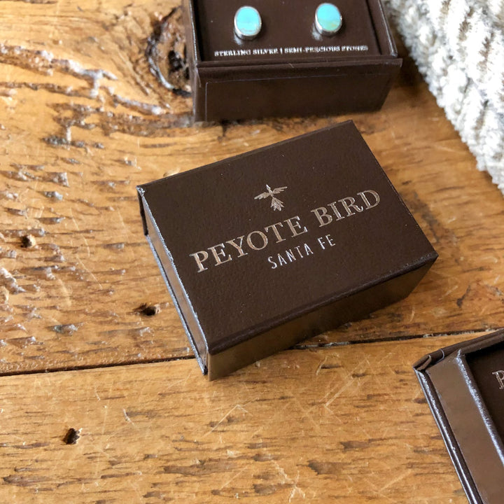A small brown box with Savannah Sevens western life{&}style and a bird logo in silver on the lid, placed on a rustic wooden surface near other jewelry boxes, perfect for storing Falcon Earrings.