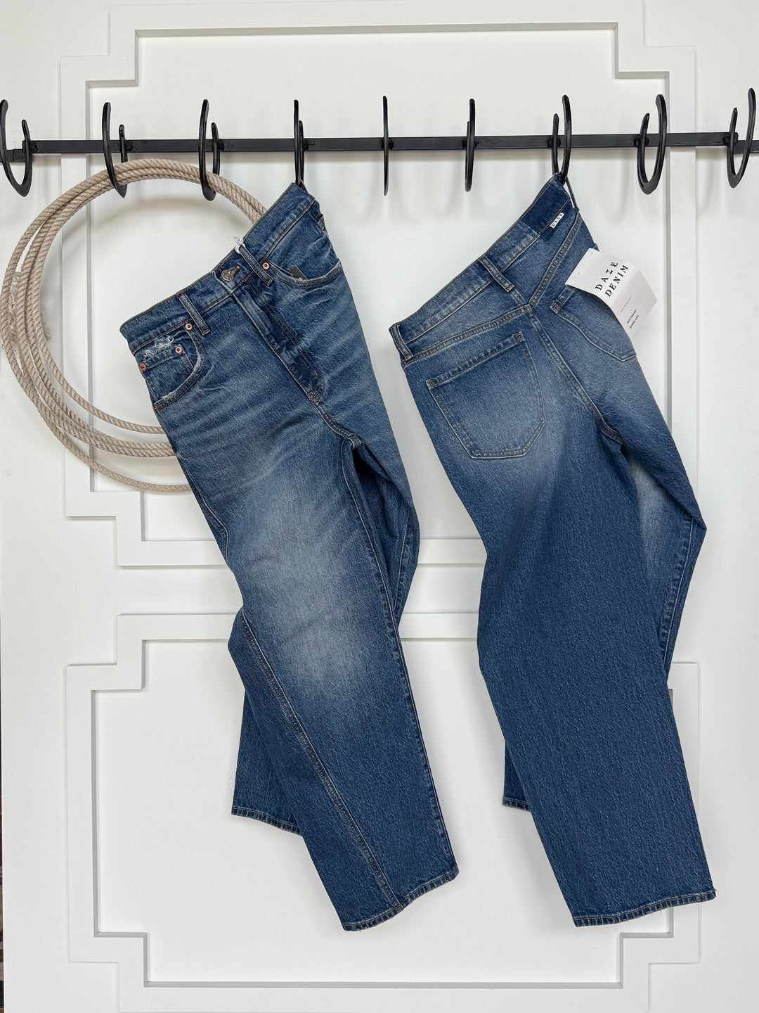 Two pairs of Savannah Sevens western life{&}style's The McKinney High Rise Barrel Jean hang by their belt loops on black hooks against a white paneled wall. A coiled rope hangs to the left, and a price tag is attached to the right pair.