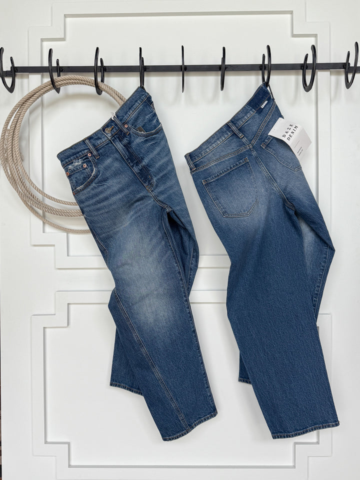 Two pairs of Savannah Sevens western life{&}style's The McKinney High Rise Barrel Jean hang by their belt loops on black hooks against a white paneled wall. A coiled rope hangs to the left, and a price tag is attached to the right pair.