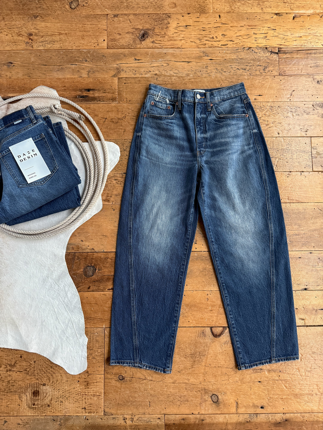 The McKinney High Rise Barrel Jean in dark blue by Savannah Sevens western life{&}style is displayed on a wooden floor beside folded medium wash denim and a white textured cloth.