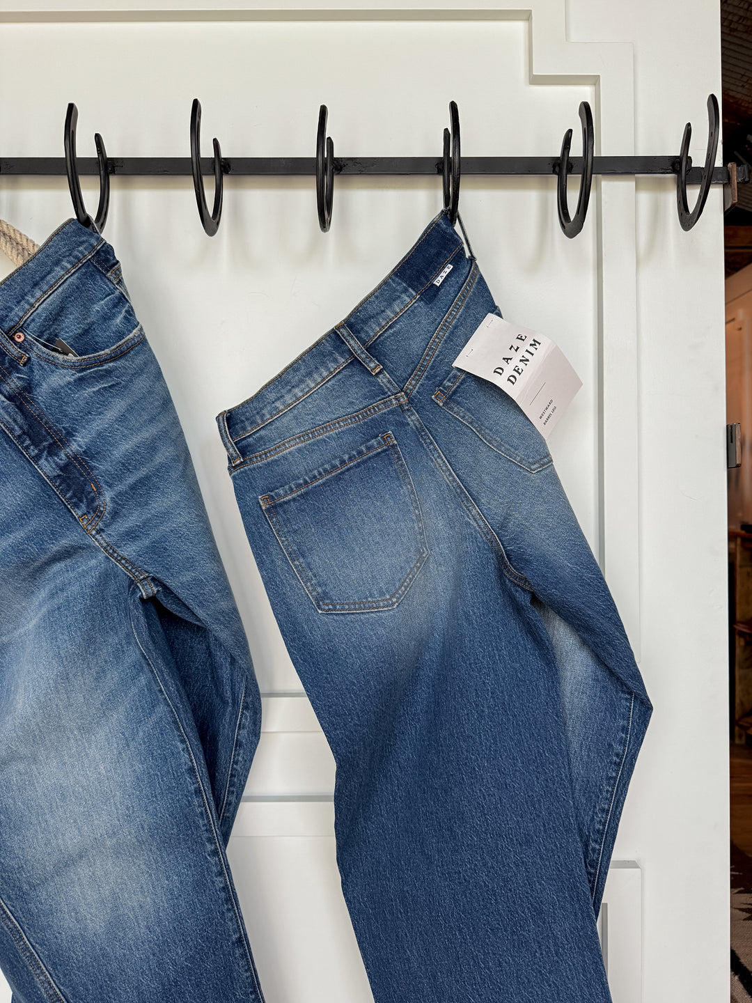 Two pairs of The McKinney High Rise Barrel Jean by Savannah Sevens western life{&}style hang on black hooks against a white wall, featuring classic stitching, a medium faded wash, and one with a visible DAYA DENIM tag in the back pocket.