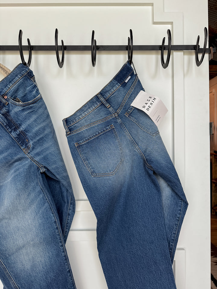 Two pairs of The McKinney High Rise Barrel Jean by Savannah Sevens western life{&}style hang on black hooks against a white wall, featuring classic stitching, a medium faded wash, and one with a visible DAYA DENIM tag in the back pocket.