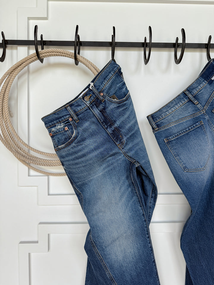 A pair of medium wash Savannah Sevens western life{&}style The McKinney High Rise Barrel Jeans hangs on a black metal hook rack mounted on a white wall, with a coil of rope and another pair of barrel jeans partially visible to the right.