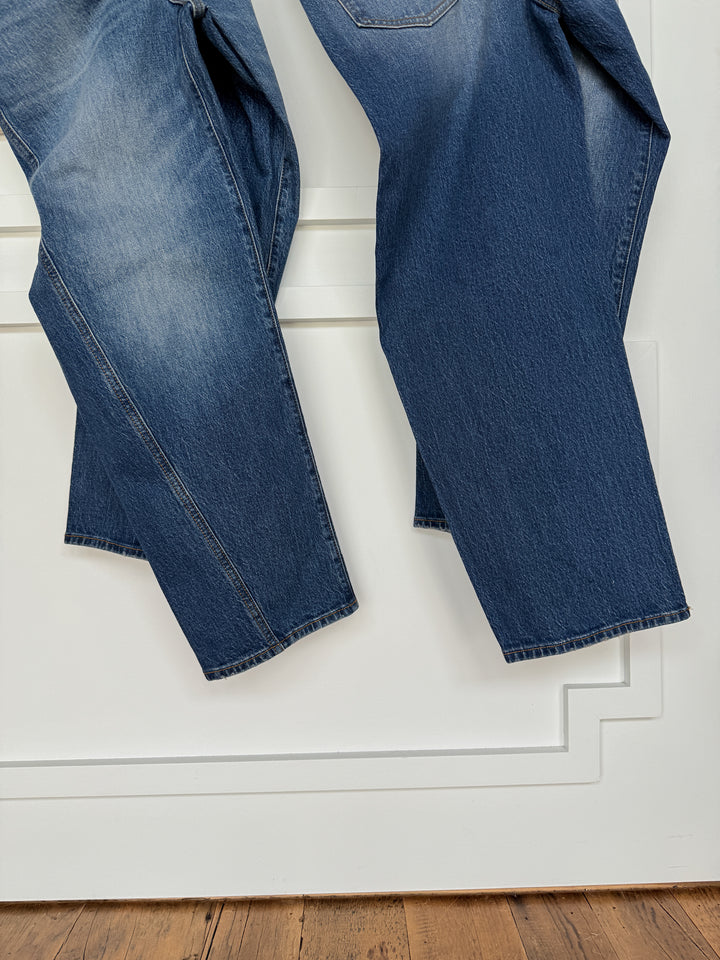 Two pairs of The McKinney High Rise Barrel Jean from Savannah Sevens western life{&}style, in medium wash with subtle fading and visible texture, hang side by side in front of a white wall above a wooden floor.