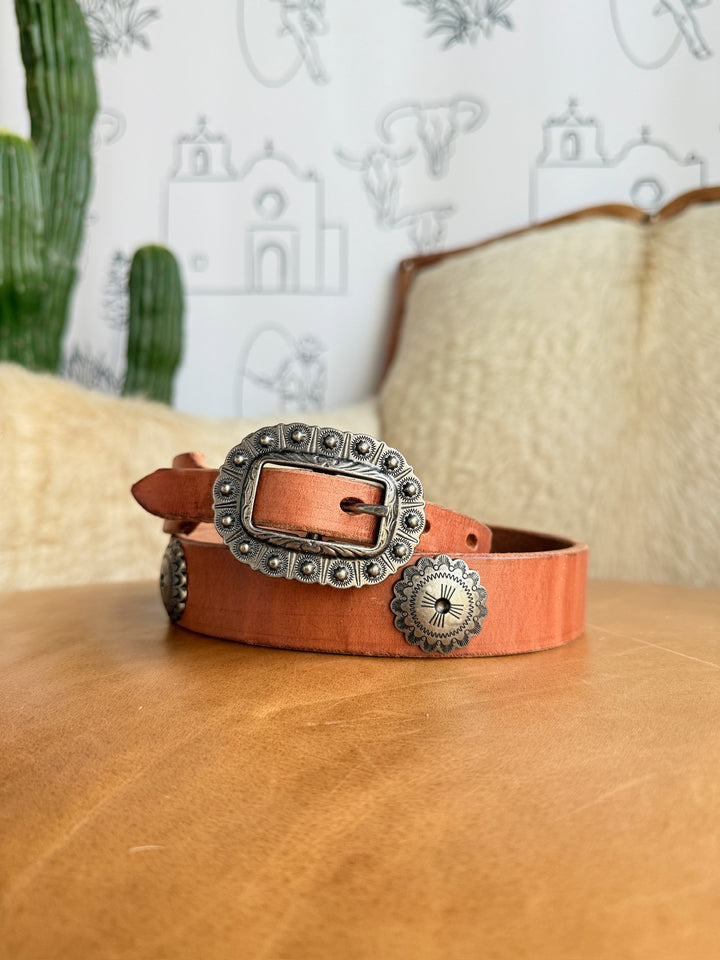 The Monticello Belt by Savannah Sevens western life{&}style, featuring cognac leather, southwest silver conchos, and an interchangeable buckle, is coiled on a tan surface with a cactus and light furniture against a line art wall.