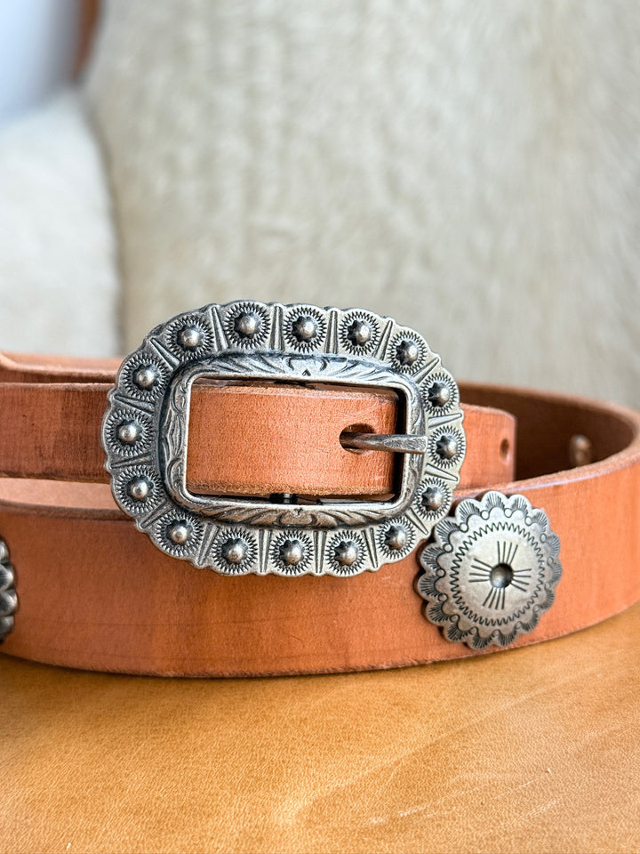 A close-up of the Monticello Belt by Savannah Sevens western life{&}style, featuring cognac leather, a large ornate interchangeable buckle, and southwest-inspired silver conchos on a light textured background.