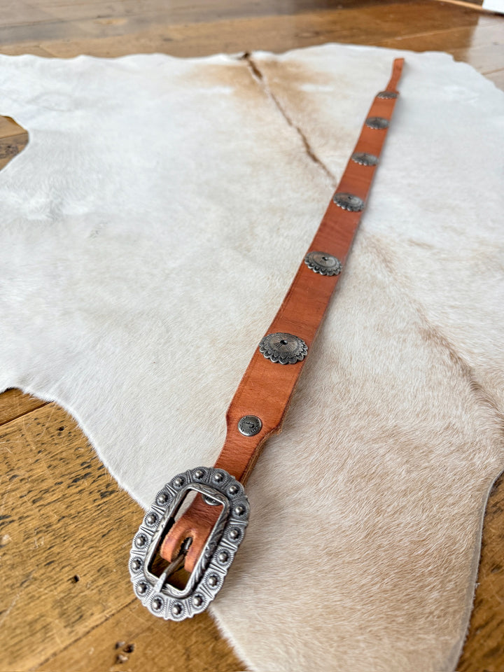 The Monticello Belt by Savannah Sevens western life{&}style features cognac leather with southwest silver conchos and a large engraved interchangeable buckle, shown on animal hide over a wooden floor.