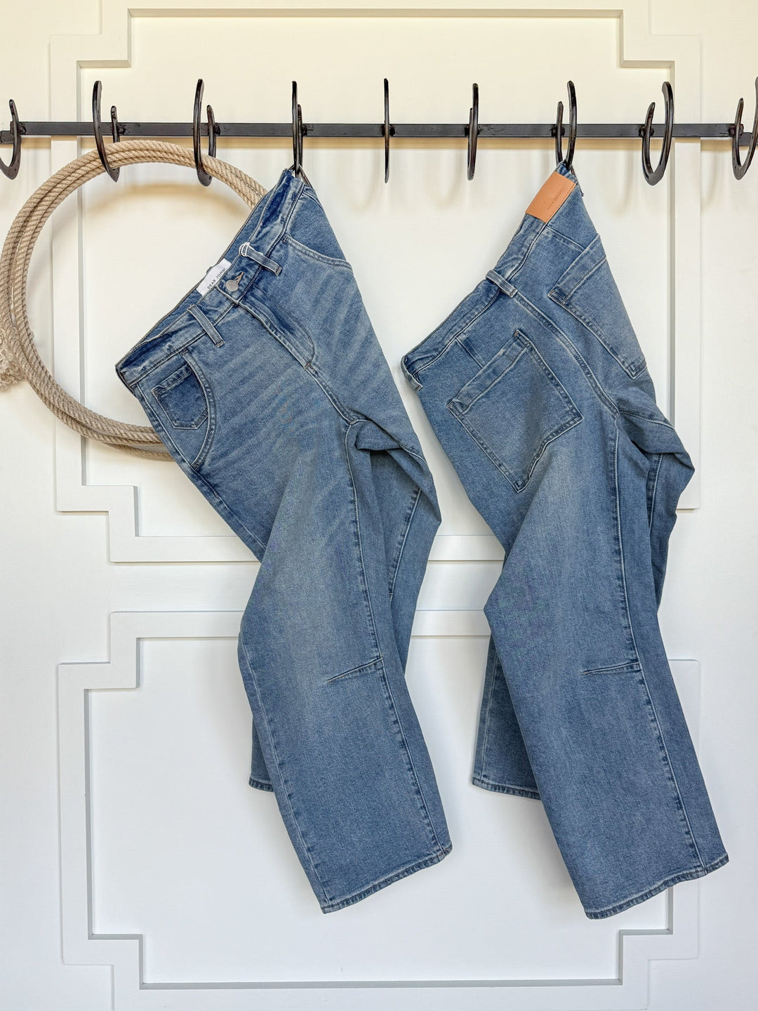 Two pairs of The Hawkin High Rise Barrel Jeans by Savannah Sevens western life{&}style hang on black hooks attached to a white panel door, with a rope on one hook and wooden floors visible below.