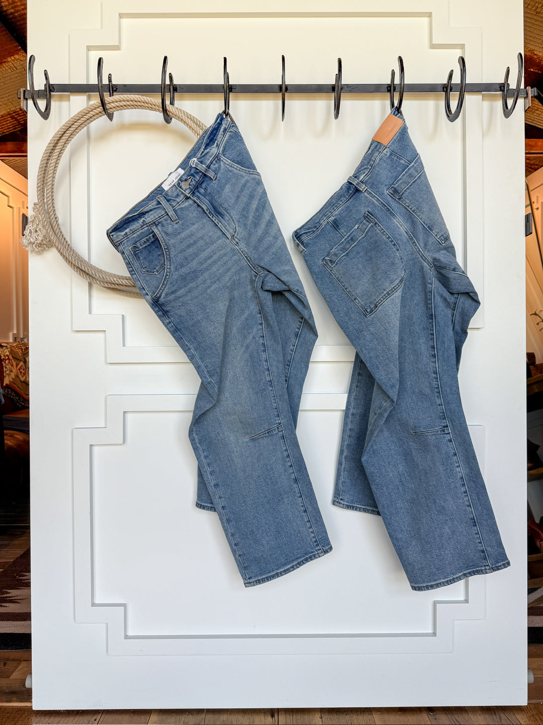Two pairs of The Hawkin High Rise Barrel Jean by Savannah Sevens western life{&}style, in blue and medium wash denim, hang on black hooks attached to a white paneled door, with wood flooring visible in the background.
