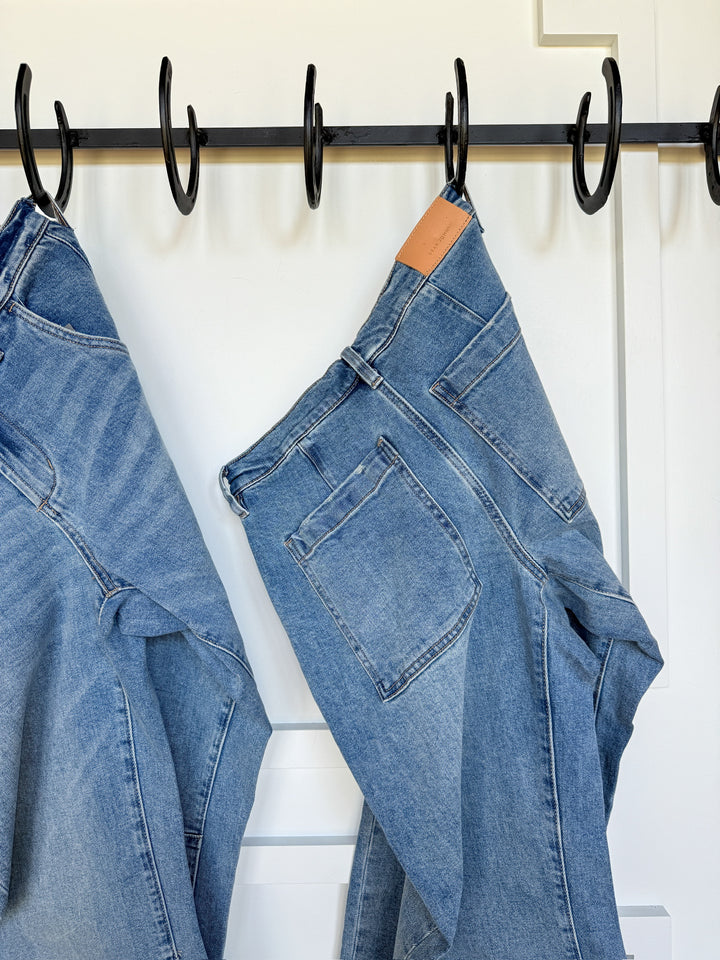 Two pairs of The Hawkin High Rise Barrel Jean by Savannah Sevens western life{&}style in medium wash denim hang side by side on black hooks against a white wall, showing the back pockets and waistbands.