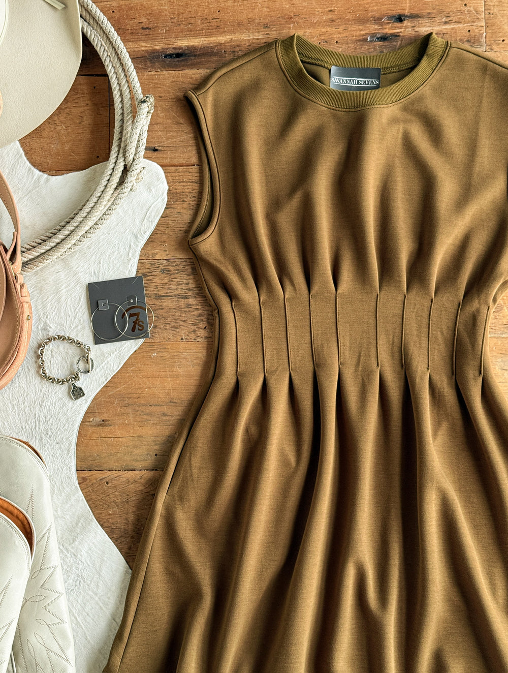 The Matewah Olive Pleat Waist Dress by Savannah Sevens western life{&}style, featuring a sleeveless bronze olive design with pleated details, is displayed on a wooden surface. It is surrounded by a white purse, gold bracelet, rope, and cowhide rug, with a gray tag attached to the dress.