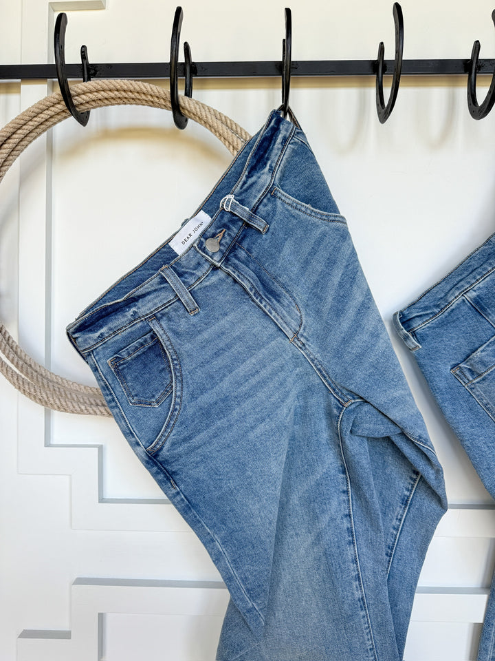The Hawkin High Rise Barrel Jean by Savannah Sevens western life{&}style in medium wash denim hangs by the belt loop on a black hook against a white wall with decorative trim and a thick rope accent.