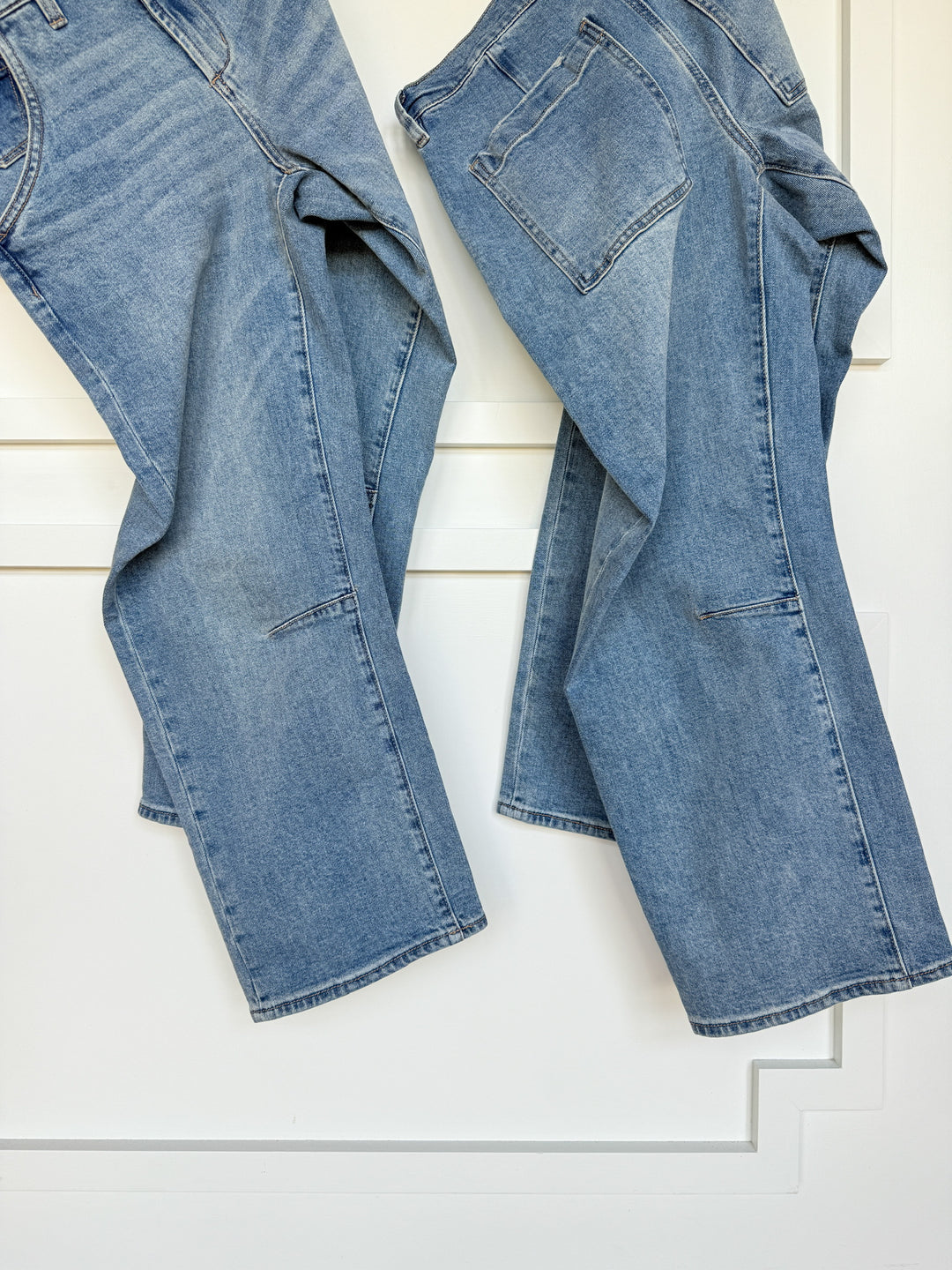 Two pairs of The Hawkin High Rise Barrel Jeans from Savannah Sevens western life{&}style hang side by side against a white wall. Both have a relaxed fit, medium wash, visible pockets, and overlapping legs that are slightly bent at the bottom.