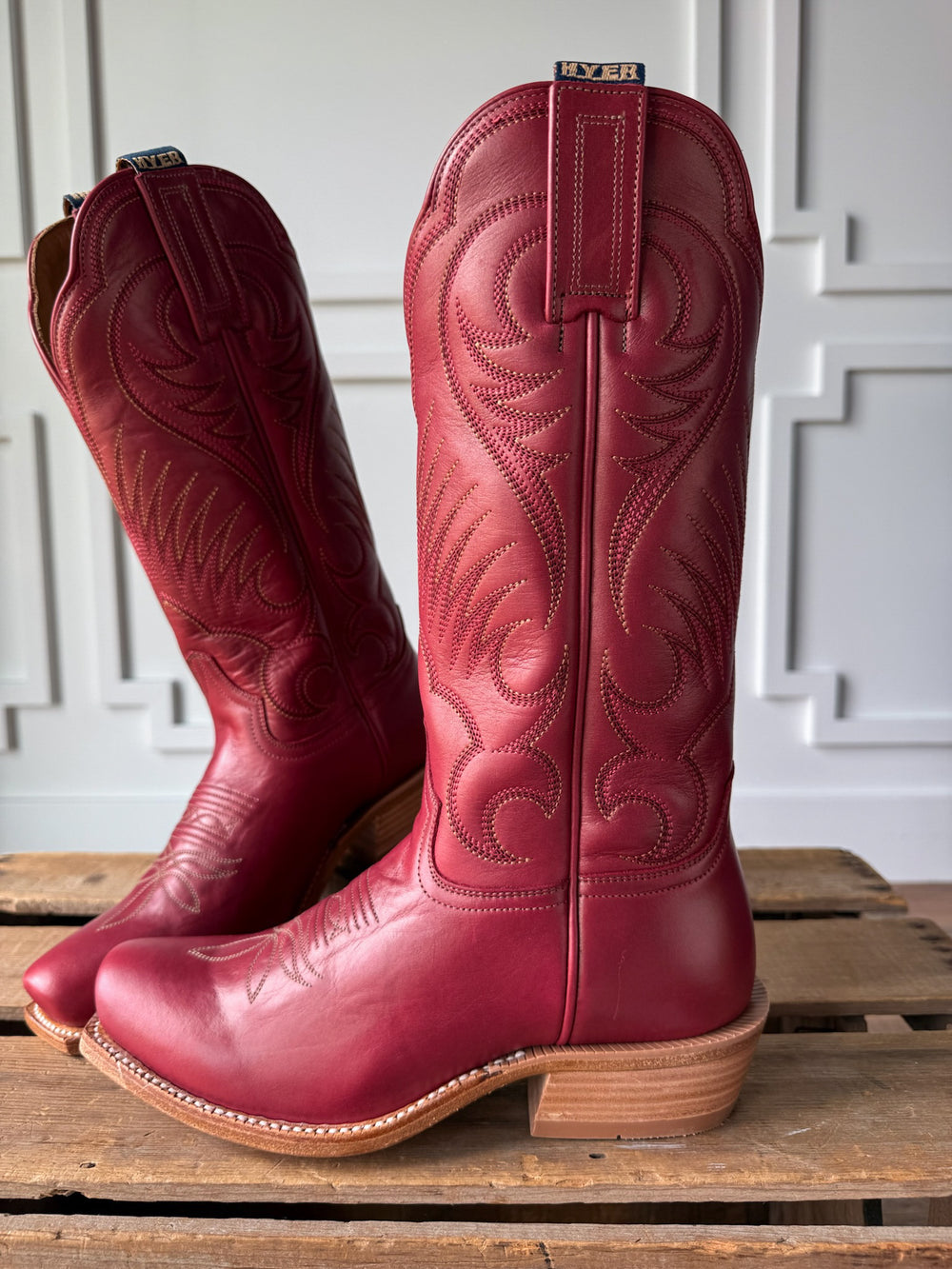 A pair of Leawood Red Western Leather Boots by Savannah Sevens western life{&}style, featuring intricate stitching, sits on a wooden pallet in front of a white, decorative wall. Boots by Hyer.