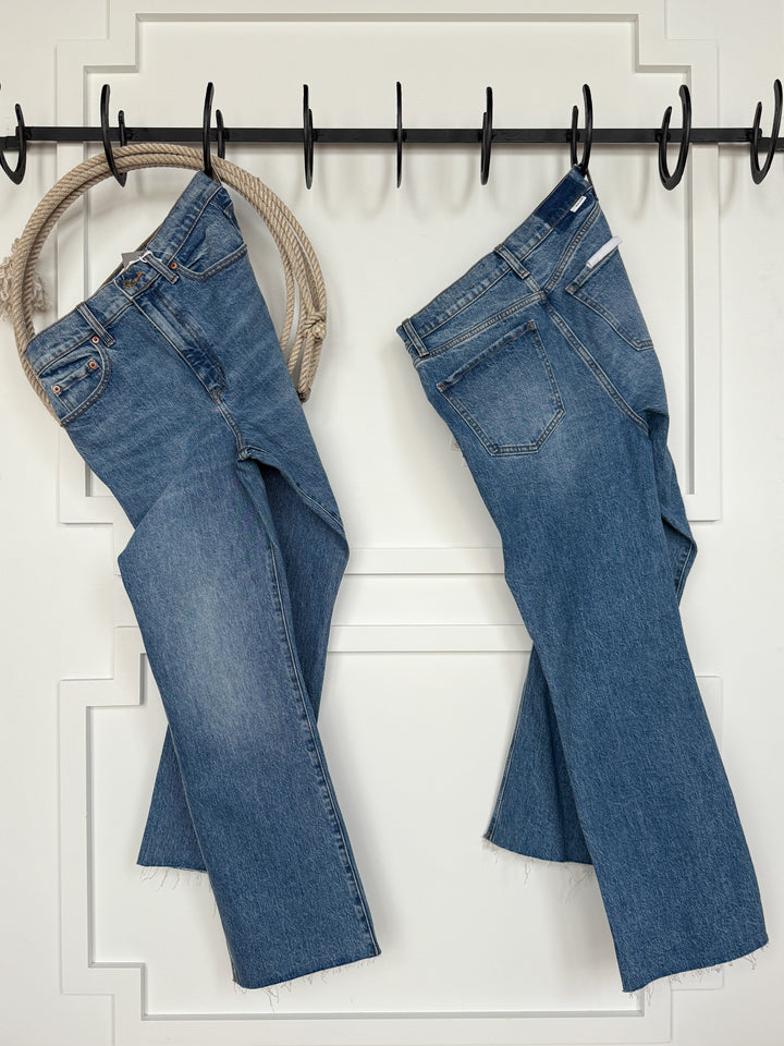Two pairs of medium wash jeans hang by their belt loops on black hooks mounted to a white paneled wall above wood floors. The Nolan High Rise Ankle Jean by Savannah Sevens western life{&}style features a raw cut hem. A rope loop also hangs from the hooks.