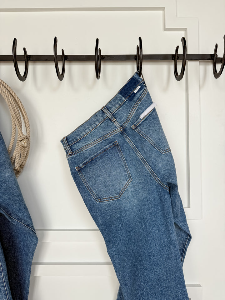 The Nolan High Rise Ankle Jean by Savannah Sevens western life{&}style, in medium wash with a raw cut hem, hangs by a belt loop on a black metal hook rack against a white wall beside part of a denim jacket and coiled rope.
