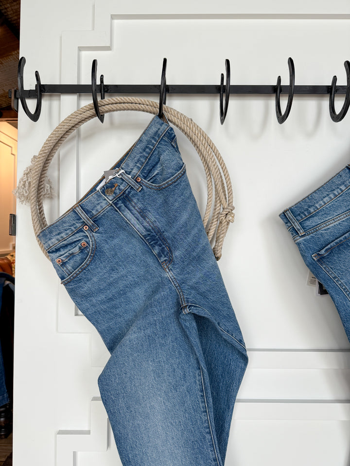 The Nolan High Rise Ankle Jean by Savannah Sevens western life{&}style hangs by its belt loops from a rope on a wall-mounted rack. Another pair is partially visible on the right, with a white paneled door in the background.