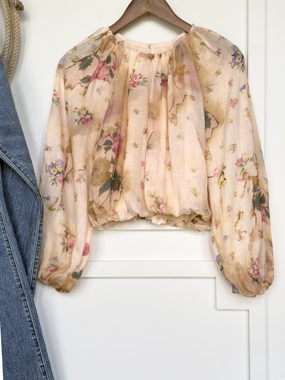 The Rosita Floral Bubble Hem Top by Savannah Sevens western life{&}style—a sheer, long-sleeve blouse with a pastel floral print, balloon sleeves, gathered neckline, and bubble hem—hangs on a wooden hanger next to a blue denim jacket. Its loose, billowy fit showcases its feminine style.