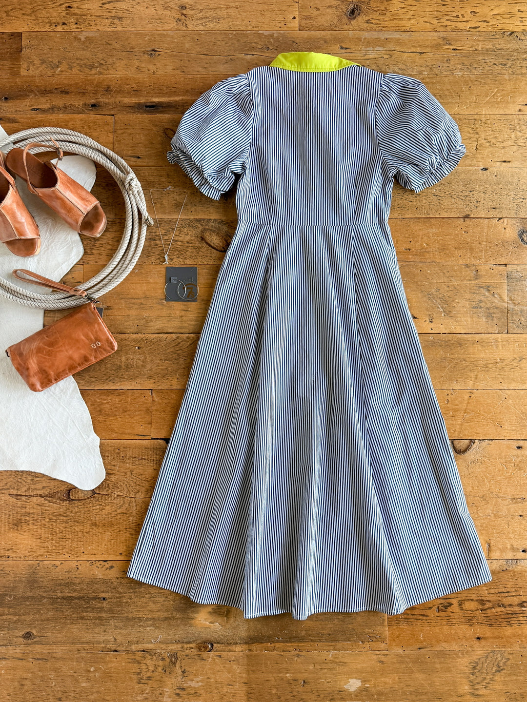 The Tererro by Savannah Sevens western life{&}style, a blue and white cotton midi dress with puffed sleeves and a yellow collar, is laid flat on a wooden floor with tan sandals, a coiled rope, and a small brown bag.
