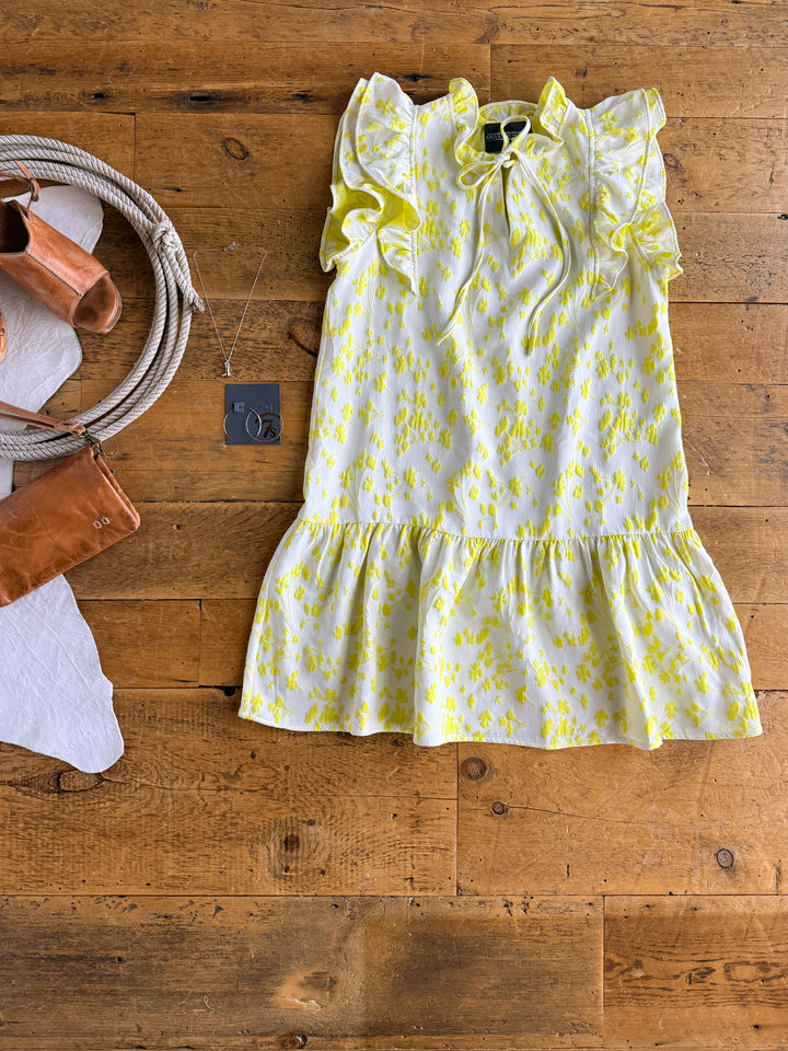 The Spring Valley dress by Savannah Sevens western life{&}style, featuring a yellow and white lemon floral pattern with ruffles, is displayed on a wooden floor with tan sandals, a small brown purse, and a silver necklace.