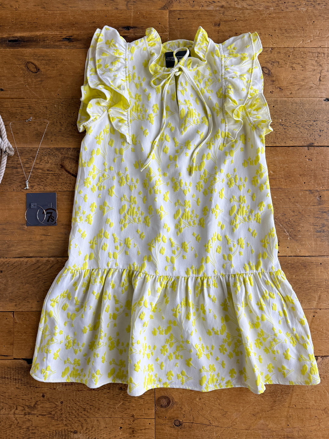 The Spring Valley dress by Savannah Sevens western life{&}style, featuring short sleeves and layered ruffle shoulders in an electric lemon floral print on white, is laid flat with silver hoop earrings and a necklace beside it.