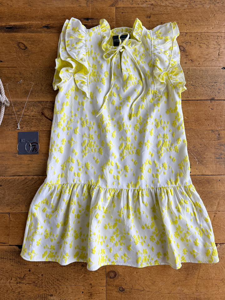 The Spring Valley dress by Savannah Sevens western life{&}style, featuring short sleeves and layered ruffle shoulders in an electric lemon floral print on white, is laid flat with silver hoop earrings and a necklace beside it.