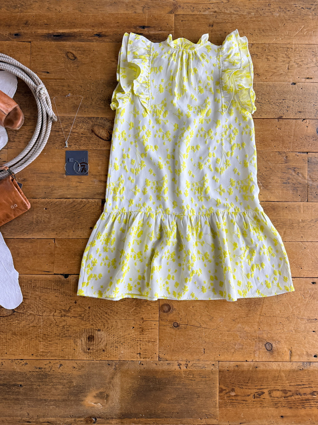 The Spring Valley dress by Savannah Sevens western life{&}style—a yellow and white electric lemon floral piece with layered ruffle shoulders—is laid flat on wood, next to a brown leather bag and hoop earrings.