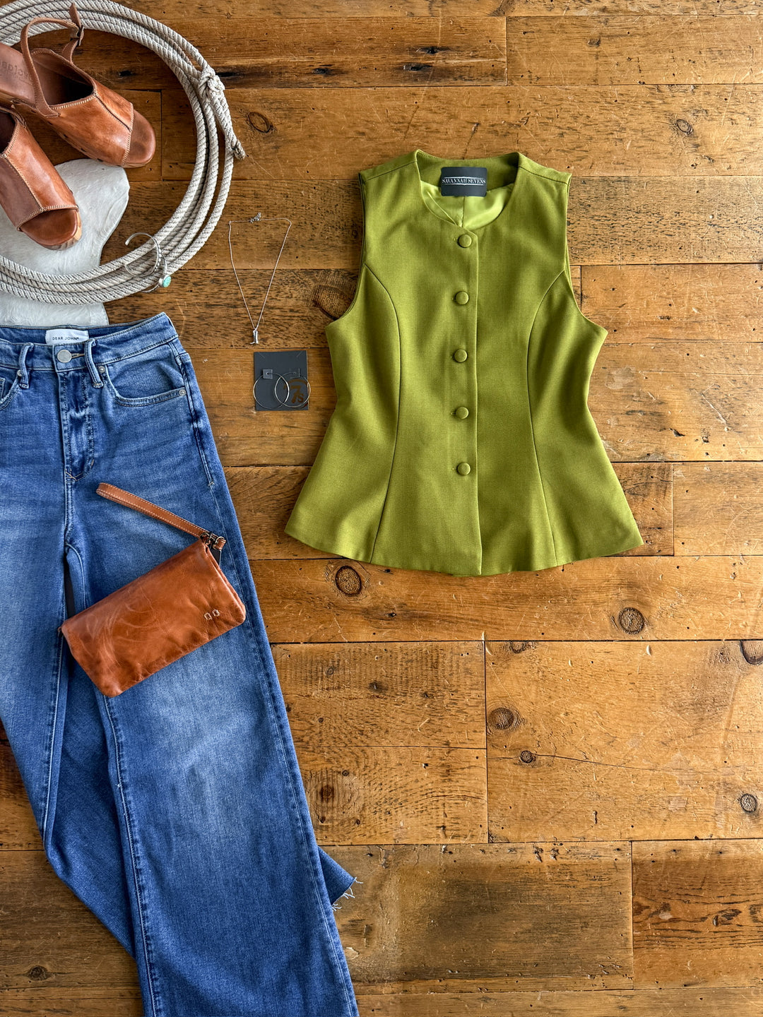 A flat lay features The Pamona blue jeans by Savannah Sevens western life{&}style, styled with an olive sleeveless tailored button-up, brown heeled sandals, a brown wristlet, necklace, and coiled rope on a wooden surface.