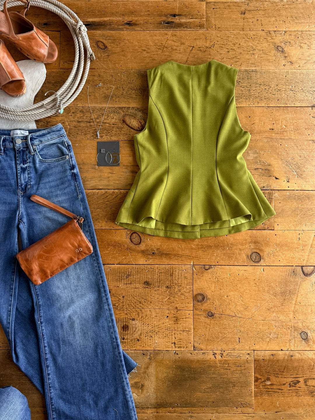 The Pamona by Savannah Sevens western life{&}style—a tailored olive sleeveless top—is laid flat on a wooden floor with blue jeans, a brown leather clutch, tan sandals, a silver necklace, and hoop earrings.
