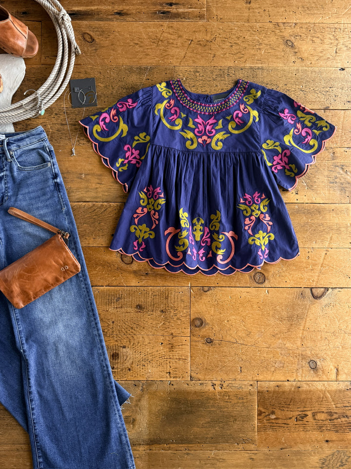 The Montevello by Savannah Sevens western life{&}style is a navy flutter sleeve blouse with pink, yellow, and green floral embroidery, shown on a wooden floor with blue jeans, a brown leather bag, and a white hat for casual charm.