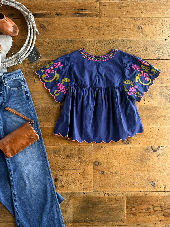 The Montevello by Savannah Sevens western life{&}style, a blue floral embroidered top with colorful sleeve patterns, is displayed on a wooden floor beside blue jeans and a brown leather purse.