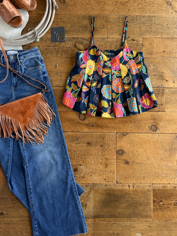 Flat lay of The Pacifica petite floral top by Savannah Sevens western life{&}style, blue jeans, brown fringe purse, tan sandals, white sun hat, silver necklace, and hoop earrings on a wooden floor.