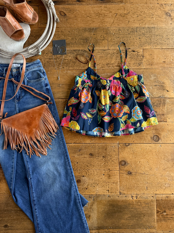 A flat lay of The Pacifica floral sleeveless top by Savannah Sevens western life{&}style, paired with blue jeans, a tan fringe crossbody bag, brown heeled sandals, a silver necklace, and earrings on a wooden floor.