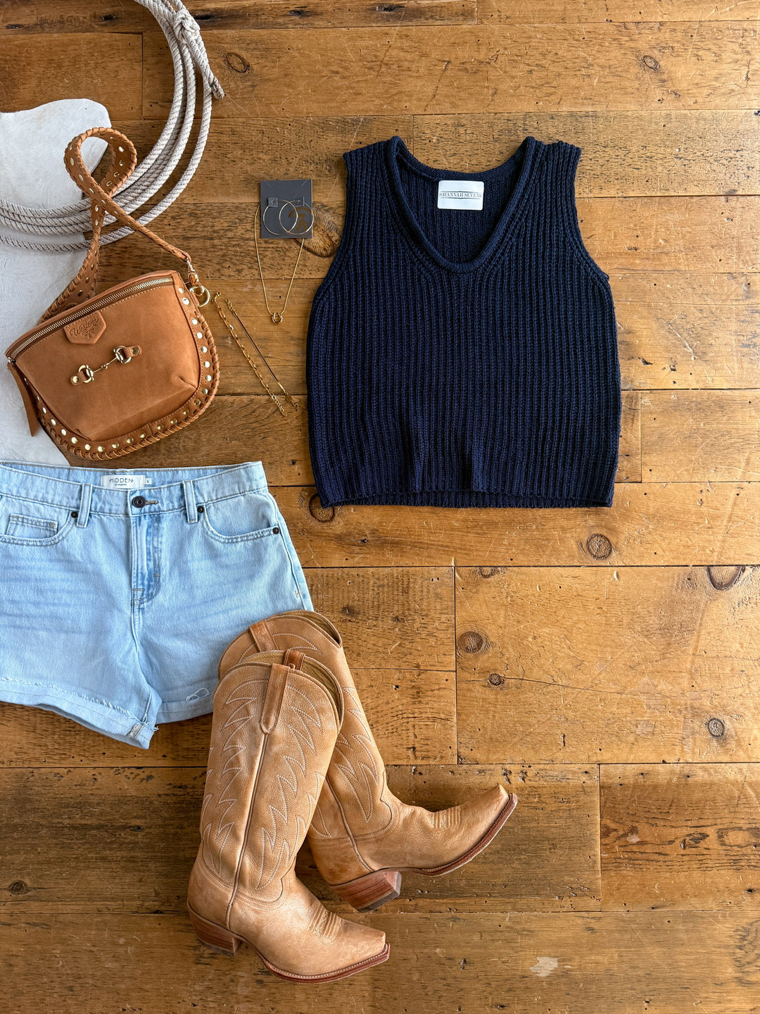 Flat lay of The Talco sleeveless navy knit top by Savannah Sevens western life{&}style, paired with light denim shorts, tan cowboy boots, a small tan studded crossbody bag, hoop earrings, and a necklace on a wooden floor.