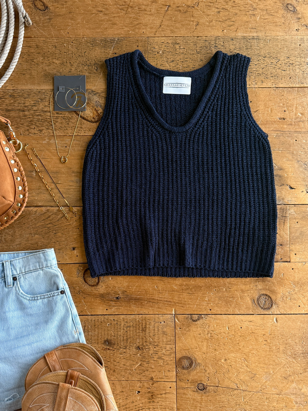 The Talco by Savannah Sevens western life{&}style, a navy knit top with a relaxed fit, is laid flat on wood alongside light denim shorts, jewelry, a tan round crossbody bag, and a brown leather purse.
