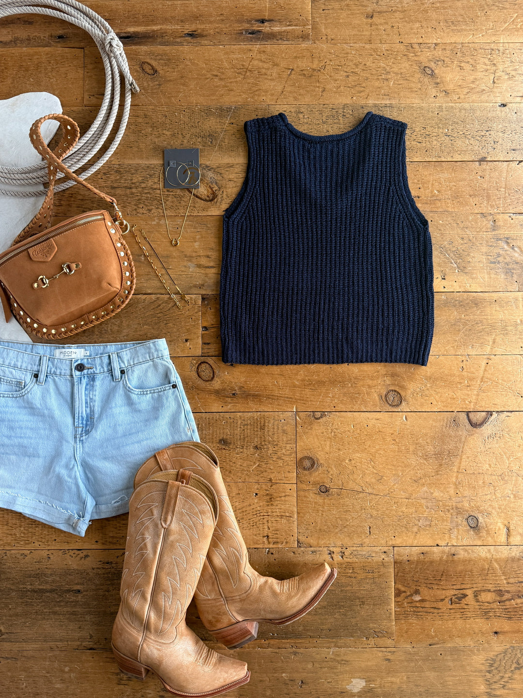 A flat lay featuring The Talco by Savannah Sevens western life{&}style: light denim shorts, a relaxed fit navy knit top, tan cowboy boots, a tan crossbody bag, and gold jewelry arranged on a wooden floor.