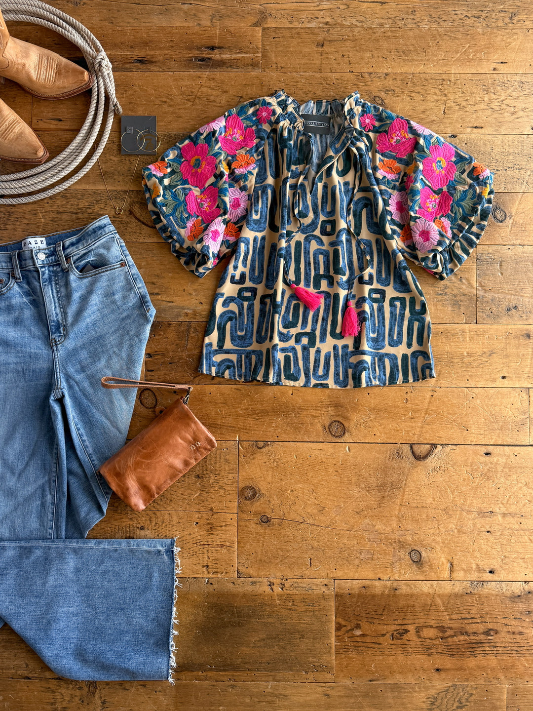 Flat lay of The Flora by Savannah Sevens western life{&}style—a floral embroidered top with pink flowers and ruffle neckline—styled with light blue jeans, a brown leather pouch, tan shoes, coiled rope, and earrings on a wooden floor.