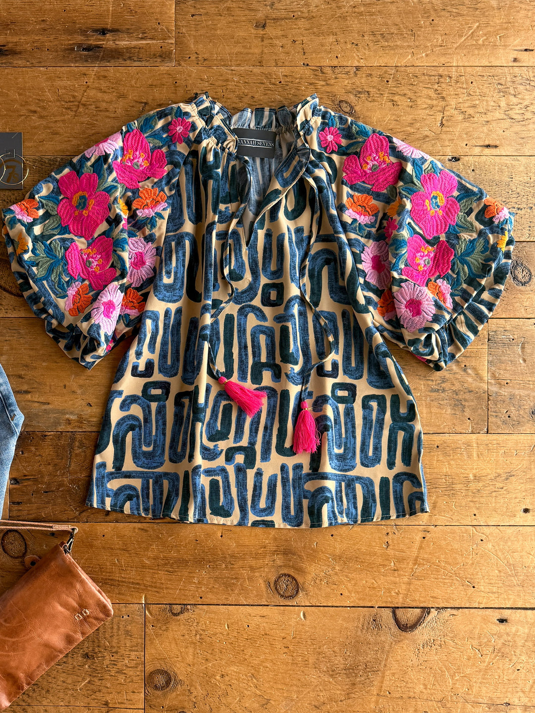 The Flora top by Savannah Sevens western life{&}style features blue geometric floral embroidery, voluminous puff sleeves, a ruffle neckline, and two pink tassels at the front, displayed flat on a wooden surface.