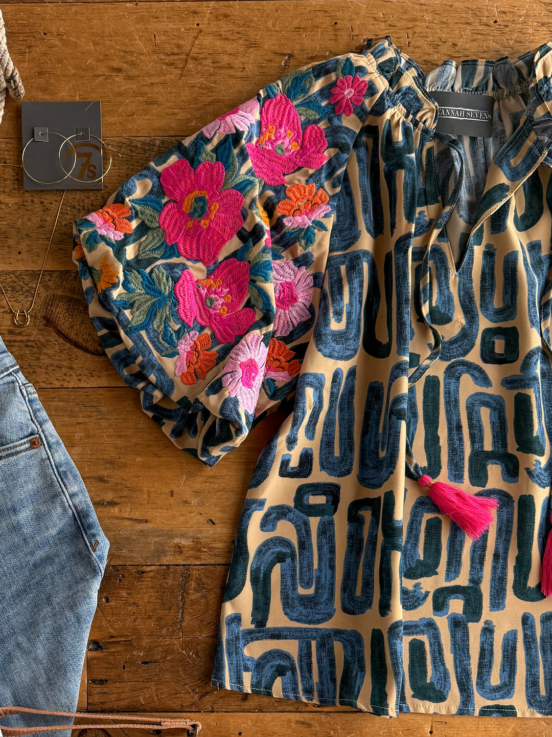 The Flora by Savannah Sevens western life{&}style: a floral embroidered top with a ruffle neckline and puff sleeves in vibrant pink and orange, styled with blue jeans, a pink tassel, and gold hoop earrings on a wooden surface.
