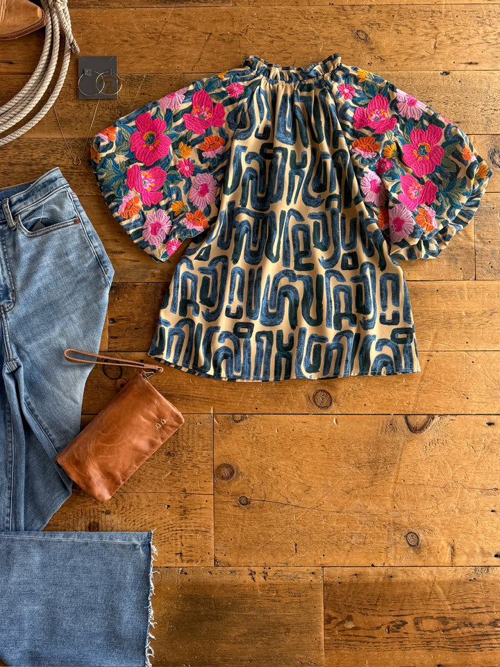The Flora by Savannah Sevens western life{&}style features bold pink, red, and orange floral puff sleeves with a blue geometric body, styled on a wooden surface beside blue jeans, a brown leather pouch, and jewelry.